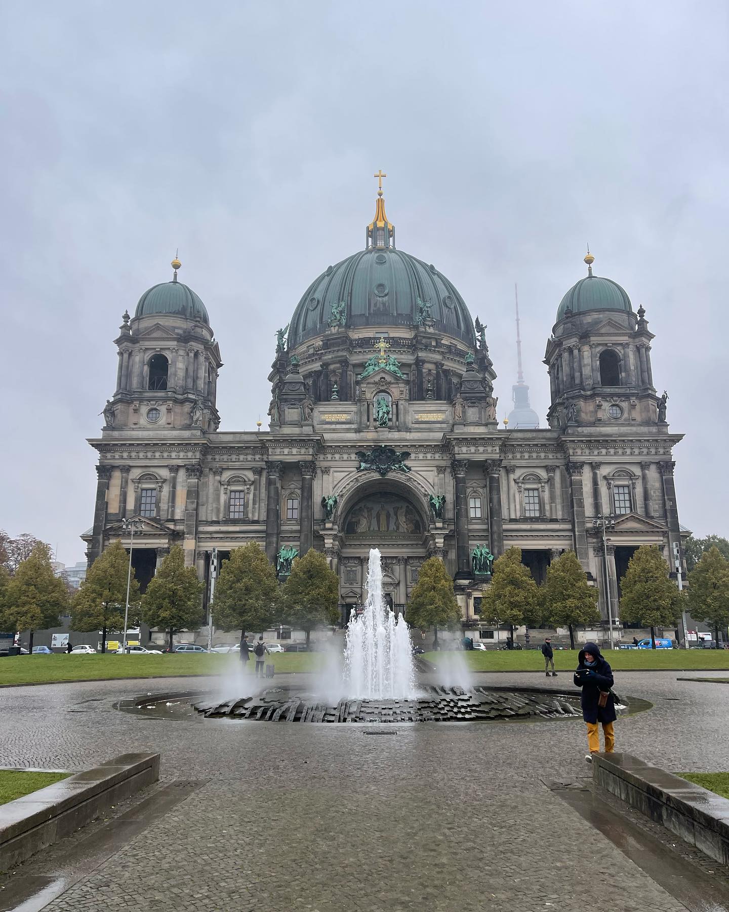Photo of the Berlin Cathedral (Berliner Dom) on an overcast day with the fountain in the foreground and a solitary person standing nearby, with the TV Tower faintly visible in the background.