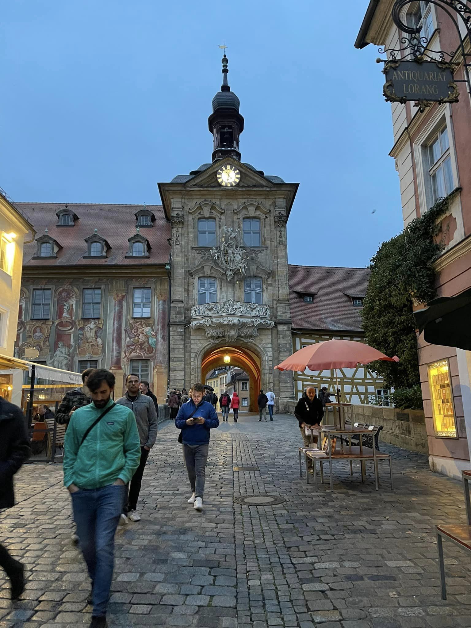 Twilight view of the Altes Rathaus with its baroque façade, ornate clock tower, and a stone archway, on a cobblestone street with pedestrians and outdoor café tables in Germany.