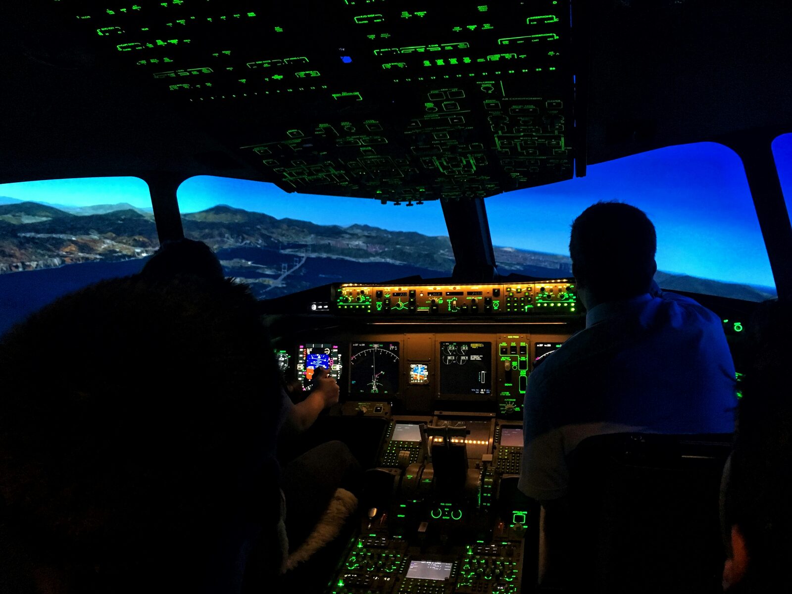 Two pilots silhouetted in a dark cockpit, the overhead panel glowing green above a blue sky