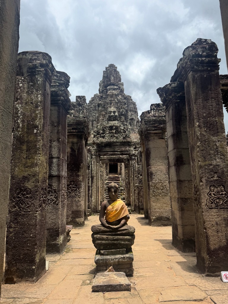 A Buddha statue draped in orange cloth, seated in a corridor with a face tower visible through the doorway beyond
