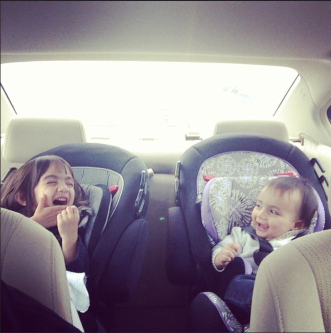 Two children in car seats in the back of a vehicle, smiling and making playful gestures. The older child on the left is laughing after making silly faces with her hands to her face, while the younger child on the right is laughing and looking at the older child.
